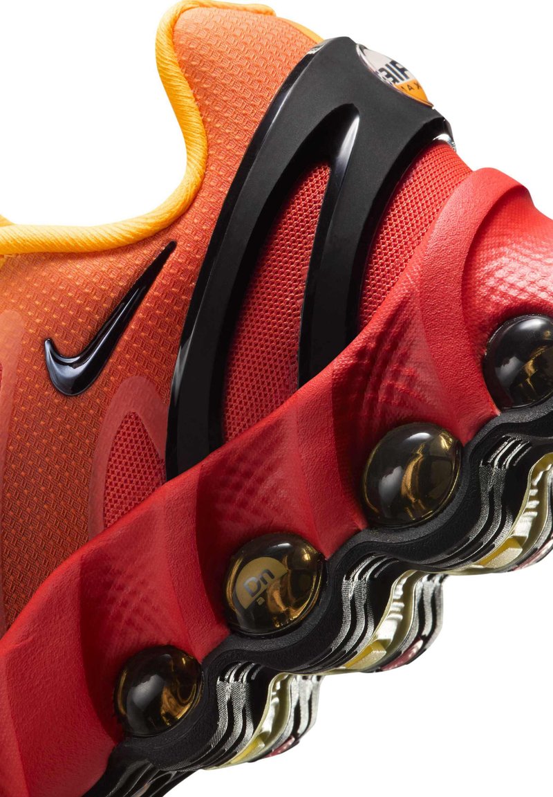 Nike cipő, amelynek narancssárga felülete textúrázott, fekete részletekkel, élénk piros talppal, amely látható légpárnás betétekkel rendelkezik, és dinamikus tervezés jellemzi.