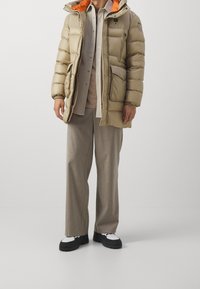 Beżowa puchowa kurtka z pomarańczową podszewką, przednimi kieszeniami i kapturem, w zestawieniu z jasnoszarą koszulą i szerokimi beżowymi spodniami. Czarno-białe buty.