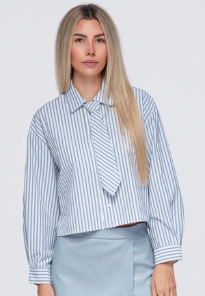 Jeune femme aux longs cheveux blonds porte un chemisier rayé bleu et blanc et une jupe bleu clair sur un fond uni.