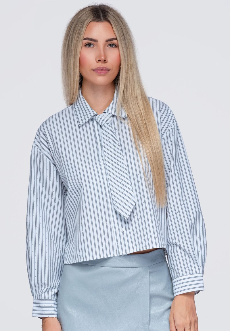 Jeune femme aux longs cheveux blonds porte un chemisier rayé bleu et blanc et une jupe bleu clair sur un fond uni.