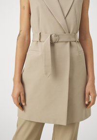 Beżowa bezrękawna marynarka z paskiem w talii zdobionym srebrnym kółkiem. Posiada dwie boczne kieszenie i gładką fakturę materiału.