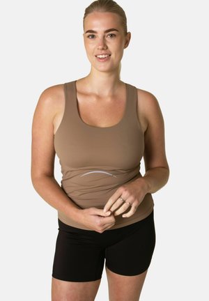 Uśmiechnięta kobieta o blond włosach, ubrana w brązowy top na ramiączkach i czarne szorty, stojąca na tle jasnego, jednolitego tła.