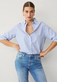 Camisa de botones a rayas azul claro y blanca con cuello y mangas enrolladas, combinada con jeans azules ajustados. Tela suave, diseño casual.