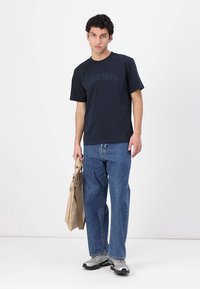 Ναυτική βαμβακερή μπλούζα με το "BELSTAFF" εκτυπωμένο στο στήθος, συνδυασμένη με ανοιχτό γαλάζιο χαλαρό τζιν παντελόνι και μπεζ τσάντα.