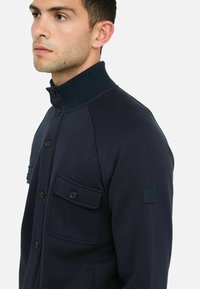 Dunkelblaues Jackett mit hohem Kragen, Knopfschluss, zwei Vordertaschen, geripptem Kragen und einem dezenten Logopatch am Ärmel.
