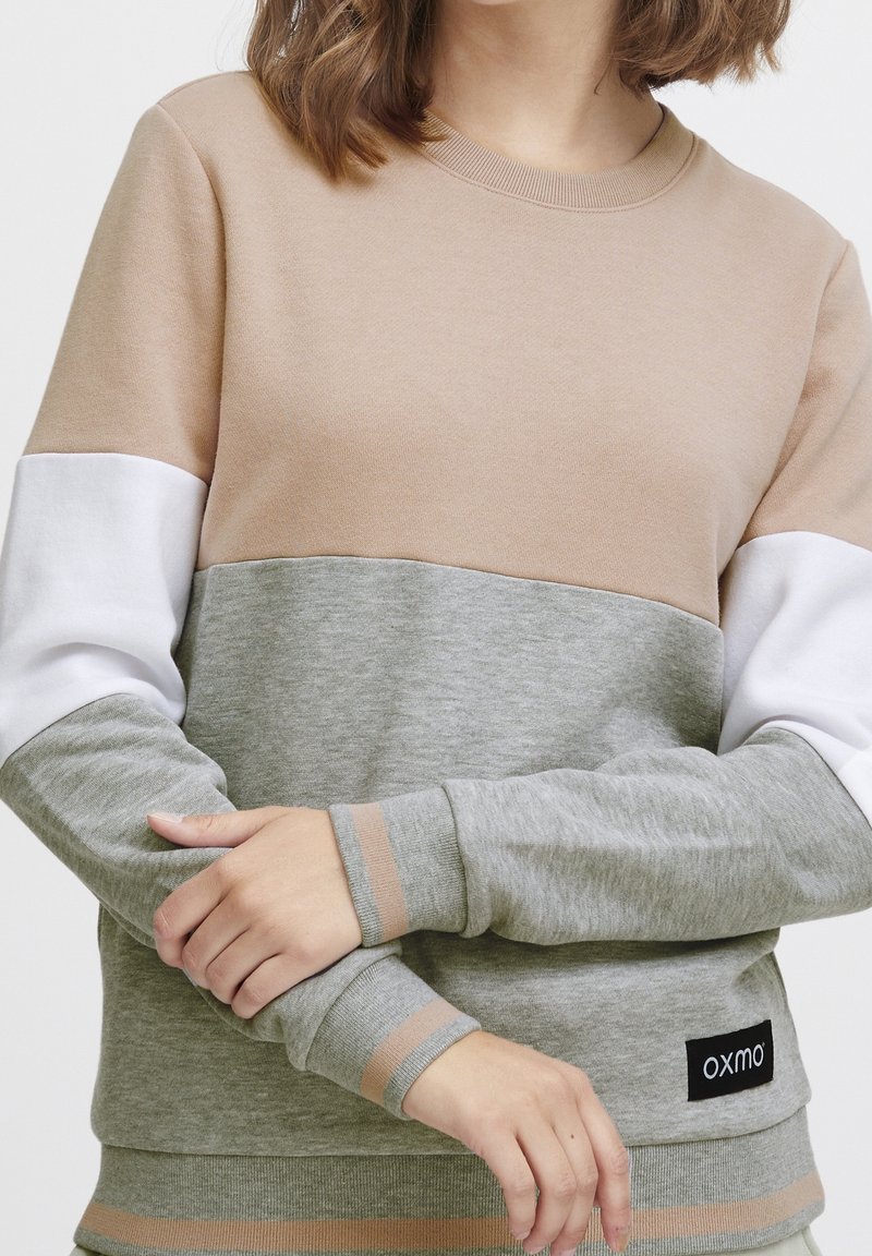 Sweatshirt beige clair et gris avec manches blanches, présentant un patch logo et des accents côtelés contrastants en beige aux poignets et à l'ourlet.