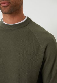 Olijfgroene trui met een gestructureerde brei-afwerking, ribbelachtige halslijn en raglanmouwen. Close-up weergave met de nadruk op het materiaal en het ontwerp.
