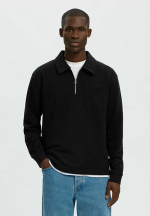 Jeune homme à la peau foncée portant une chemise noire à manches longues avec une fermeture éclair à l'avant et un jean bleu clair sur un fond uni.