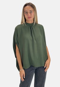 Top verde testurizzato con collo alto, maniche corte e vestibilità ampia, abbinato a jeans scuri. Presenta fessure laterali per maggiore libertà di movimento.