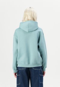 Mintgroene hoodie met een trekkoord in de capuchon, omgeslagen mouwen en een lose fit. Zachte textuur, gestikte zoom en gedragen met een donkere spijkerbroek.