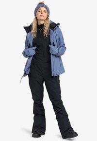Blaue isolierte Jacke mit schwarzen Latzhosen, passenden Handschuhen und einer blauen Strickmütze. Verfügt über Reißverschlüsse und Taschen für mehr Praktikabilität.