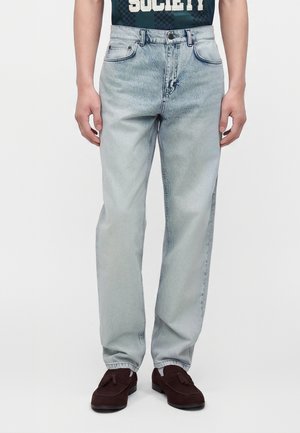 Lichtblauwe rechte spijkerbroek gedragen met bruine tassel loafers bij een staand persoon tegen een effen achtergrond.