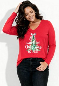 Czerwona koszula z długim rękawem i dekoltem w kształcie litery V, z białym i zielonym napisem oraz grafiką choinki z kokardą. Do tego ciemne dżinsy.