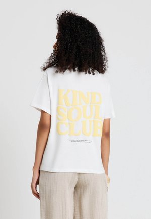 Donna con capelli ricci che indossa una maglietta bianca con una grande scritta gialla "KIND SOUL CLUB" sulla schiena e pantaloni beige chiaro.