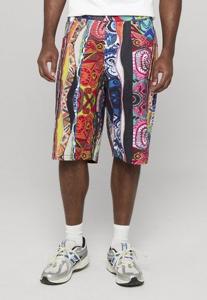 Uomo che indossa pantaloncini colorati a motivo stampato fino al ginocchio, calzini bianchi e scarpe da corsa bianche e blu su sfondo grigio.