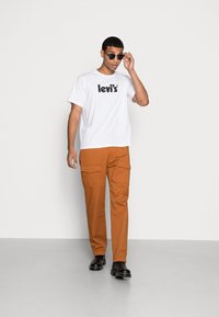 Weißes T-Shirt mit kurzen Ärmeln und schwarzem "Levi's"-Logo, kombiniert mit orangen Cargo-Hosen mit Taschen. Trägt dazu schwarze Sonnenbrillen und Stiefel.
