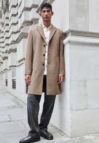 Cappotto di lana marrone con colletto a rever e bottoni frontali, abbinato a jeans neri e scarpe nere. Il cappotto ha una texture liscia e tasche laterali.