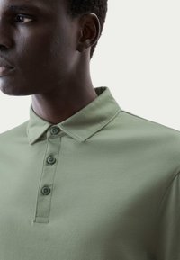 Polo tričko v tlumené zelené barvě, vyrobené z měkké tkaniny s hladkým texturem. Má límec a tři knoflíky, s přiléhavým střihem.