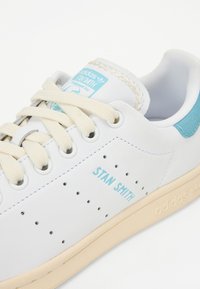 Hvide lædersneakers med lys blå accenter, perforerede detaljer, beige sål og beige snørebånd. "STAN SMITH" logo på siden.