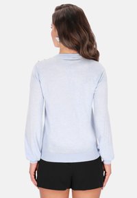 Maglione azzurro chiaro con texture morbida, maniche lunghe e orlo a coste. Presenta dettagli con bottoni lungo la linea del collo posteriore. Abbinato a pantaloncini neri.