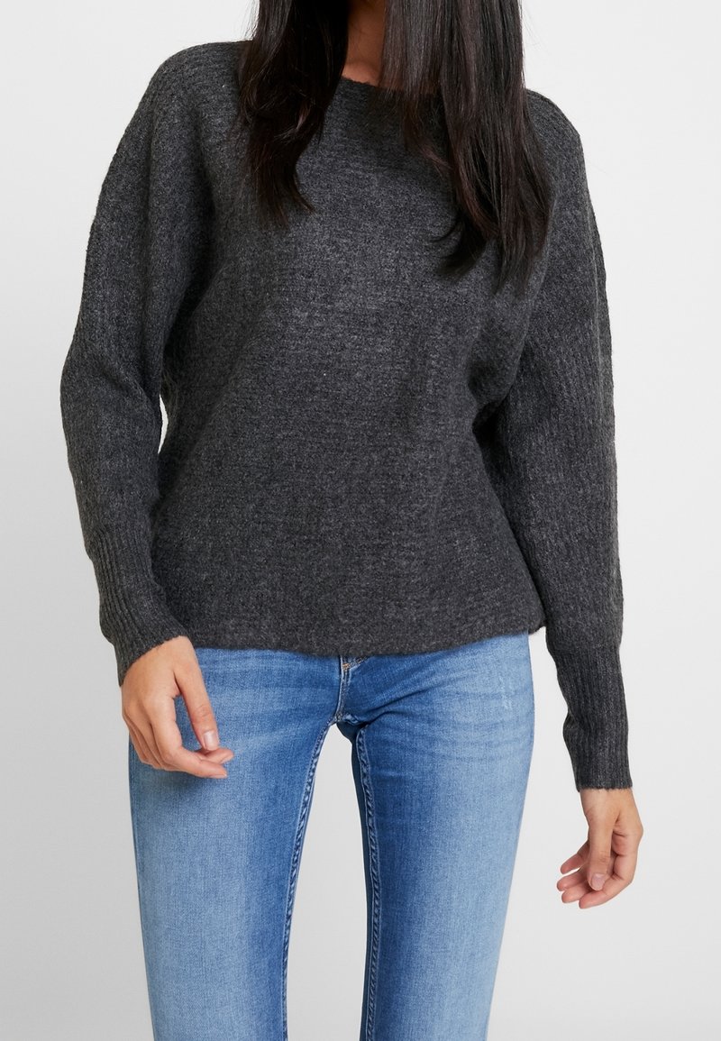 Pull tricoté gris foncé avec encolure ronde, épaules tombantes et poignets côtelés, assorti à un jean bleu clair. Texture douce.