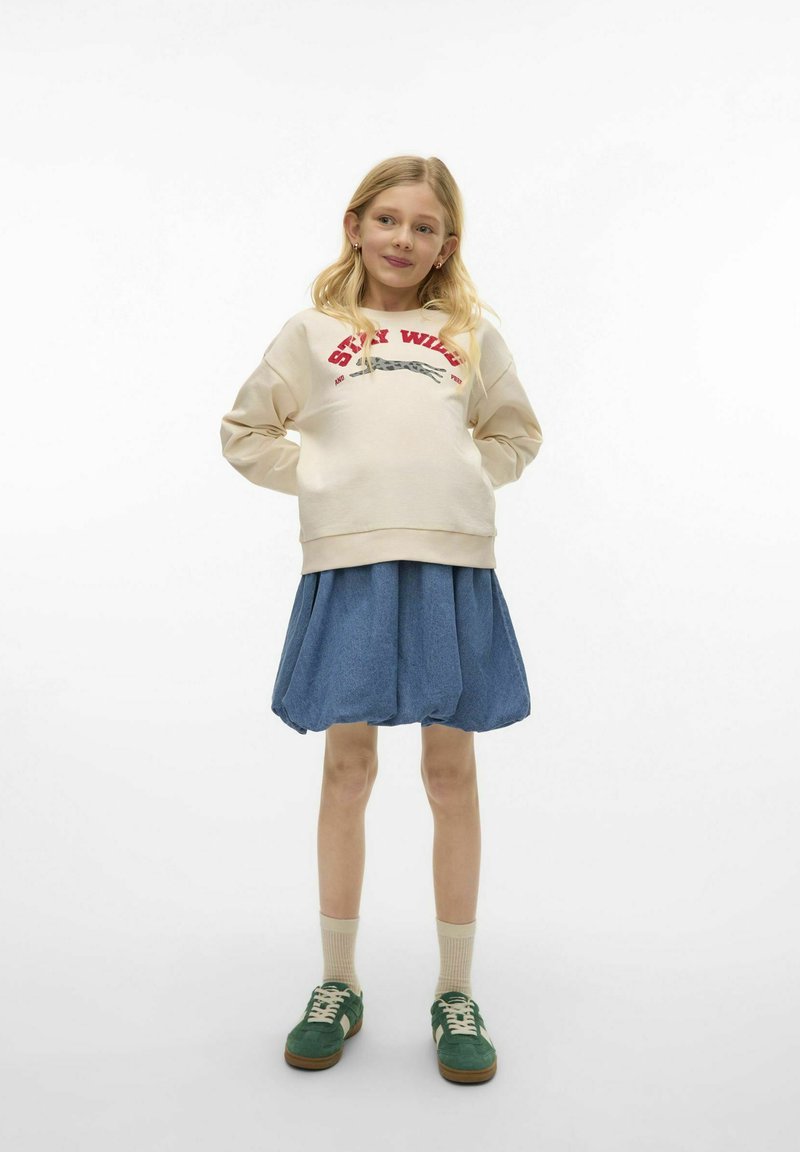 Krämsweatshirt med "Stay Wild" grafik, kombinerat med en blå denimkjol. Gröna sneakers med beige snören och korta krämfärgade strumpor.