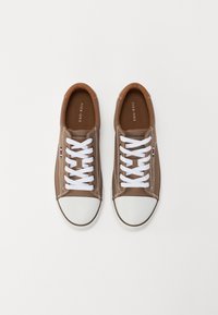 Zapatillas de lona marrón con cordones blancos, punteras de goma y costuras en contraste. Presenta una pequeña etiqueta multicolor en el lateral.