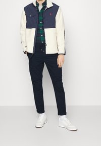 Man wearing white fleece and navy jacket, green and navy plaid shirt, navy pants, white socks, and white sneakers, standing against a plain background.