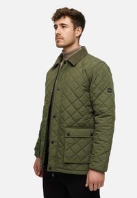 Quilted olivengrøn jakke med kordfløjskrave, trykknapper, to forlommer og sort accentlappe på ærmet. Tekstureret finish.