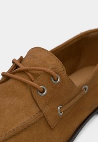 Chaussure bateau en daim marron avec des lacets tressés passant par des œillets métalliques et une couture détaillée sur la partie supérieure.