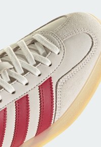 Gros plan d'une basket beige avec des lacets blancs, des bandes rouges texturées et une semelle en caoutchouc translucide brun clair.