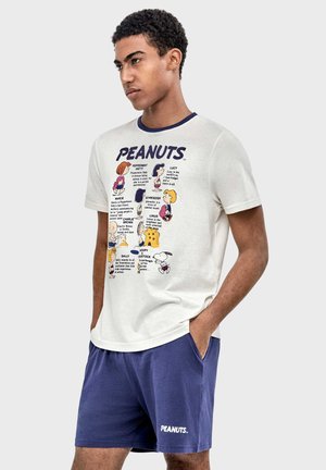 Mladić u bijeloj majici s likovima iz Peanuts serije i plavim kratkim hlačama s Peanuts logom, stoji s rukama u džepovima.