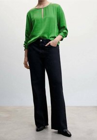 Personne portant un haut vert à manches longues avec une fente à l'avant et un pantalon noir à jambes larges, debout devant un fond clair uni.