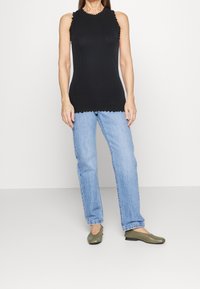 Sort ribbet tanktop uden ærmer med bølgede kanter, parret med lyseblå lige ben jeans og olivengrønne flade sko.