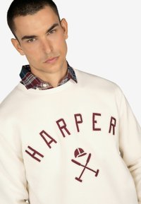 Sudadera crema con "HARPER" en bordado burdeos, superpuesta con un cuello de camisa de cuadros en tonos rojos y azules.
