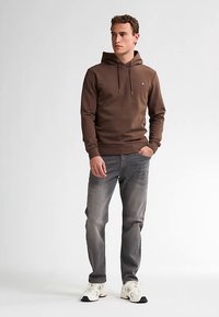 Hnědá mikina s kapucí a šňůrkami, přední kapsa ve tvaru klokánka, nošená s světle šedými džínami a bílými teniskami. Jednoduchý design, bez vzorů nebo grafiky.