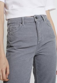 Grå kordfluweelpants med vertikal ribbad struktur, som har framfickor, knappstängning och en åtsittande midja, visad med en kort ärmad vit topp.