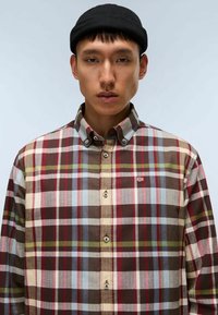 Giovane uomo con un cappellino nero e una camicia a maniche lunghe a quadri, con tonalità di rosso, marrone, blu e verde, su uno sfondo semplice.