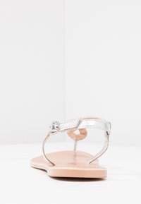 Hopeapintainen sandaali, jossa on litteä pohja, ohut nilkkavyö ja minimalistinen muotoilu. Sileä päällinen ja teksturoitu pohja.