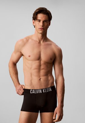Mężczyzna bez koszulki z wysportowaną sylwetką, ubrany w czarne bokserki Calvin Klein, trzyma pas jedną ręką na jednolitym szarym tle.