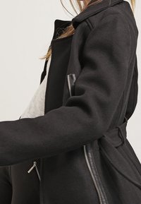 Nainen, joka pitää yllään mustaa villakangastakkia, jossa on hopeinen vetoketju ja vyötäröllä yksityiskohta, vaalean neulepaidan päällä, näkyy sivulta yksinkertaista taustaa vasten.