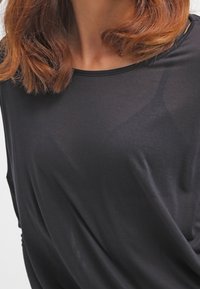 Woman with shoulder-length light brown hair wearing a sheer black top over a black camisole, shown from the neck to waist.