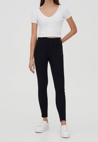 Černé úzké džíny s vysokým pasem a jemnou texturou, doplněné o bílý krátký top s vlnitým okrajem a krátkými rukávy.