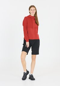 Röd zip-up jacka med strukturerad tyg, kombinerad med svarta shorts. Modellen bär svarta och grå sneakers. Enkel design, praktisk för utomhusaktiviteter.