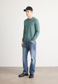 Teal sweatshirt with ribbed cuffs, paired with blue denim jeans and black leather shoes. Black cap with white logo. Simple, casual outfit.