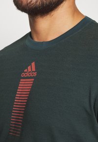Tmavě zelené sportovní tričko s kulatým výstřihem, krátkými rukávy a výrazným červeným logem Adidas s vodorovnými pruhy na levé straně.