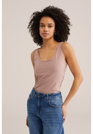 Donna con capelli ricci che indossa un top smanicato color malva chiaro e jeans blu, in piedi con una mano vicino alla tasca su sfondo semplice.