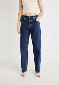 Ženska nosi visoko pasne temno modre ravne hlače, skrajšan bež top in bele sandale z odprtimi prsti ter stoji proti enobarvni ozadju.