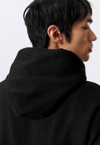Schwarzer Kapuzenpullover aus einem weichen, strukturierten Stoff, mit einer hervorgehobenen Kapuze und glatten Nähten. Minimalistisches Design ohne sichtbare Logos.