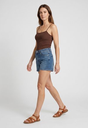 Femme marchant portant un haut marron sans manches, un short en jean bleu et des sandales plates marron sur un fond clair uni.